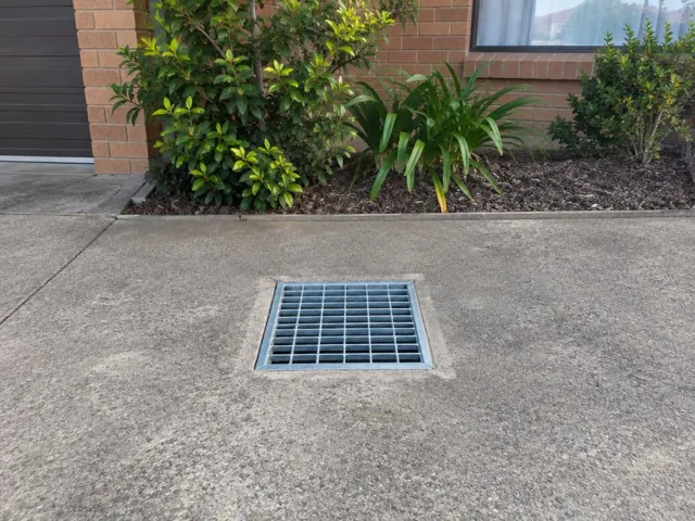 Concrete driveway with a square metal stormwater grate beside brick house walls and shrubs in Wakeley